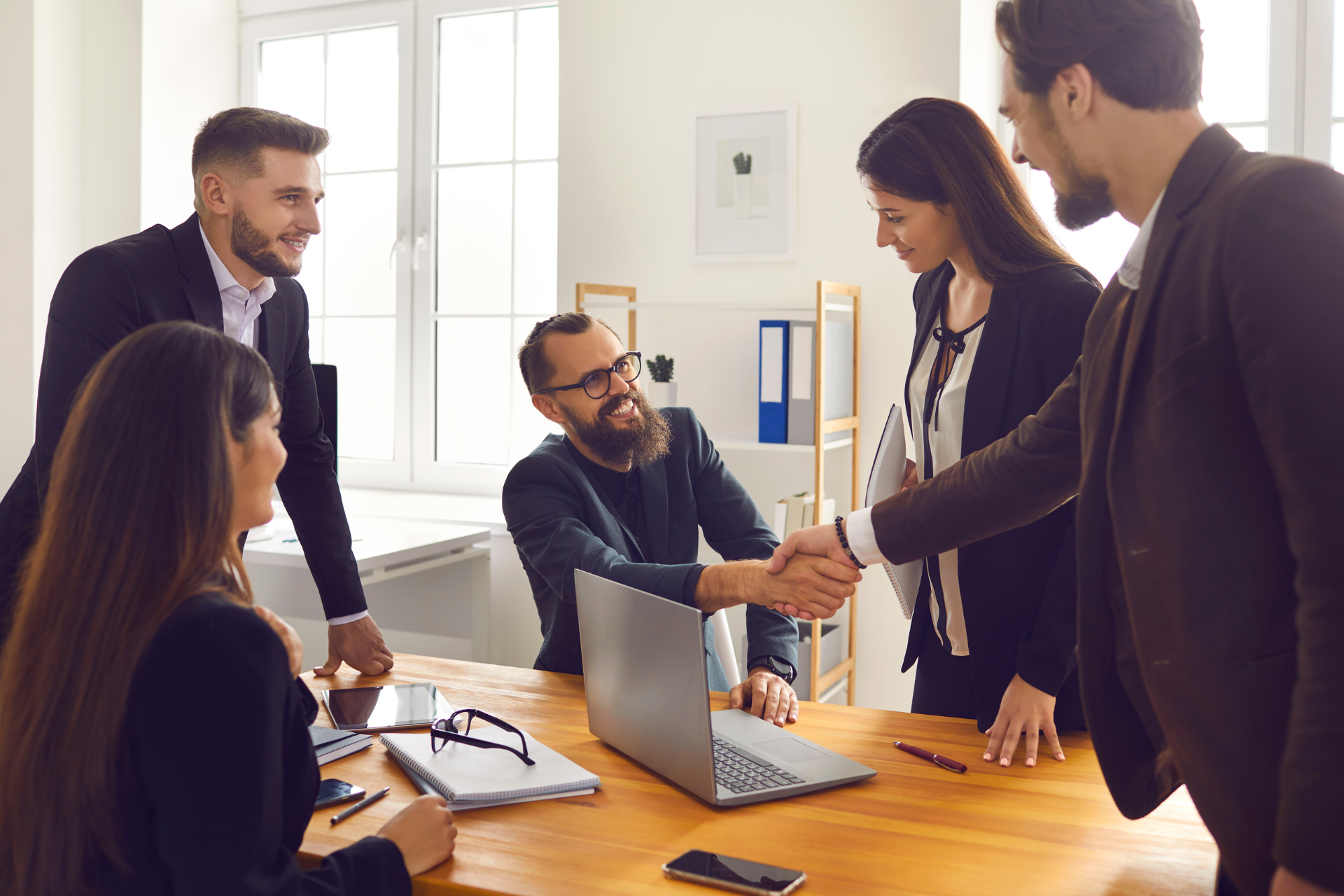 This photograph shows a group of professionals around a table and two people shaking hands. It’s a stock image so the individuals should not be identified. It’s being used to illustrate that the article it represents is about a deal being made between two businesses. The text alternative is “Should You Sell to a Competitor?” as this best describes the intended impression.
